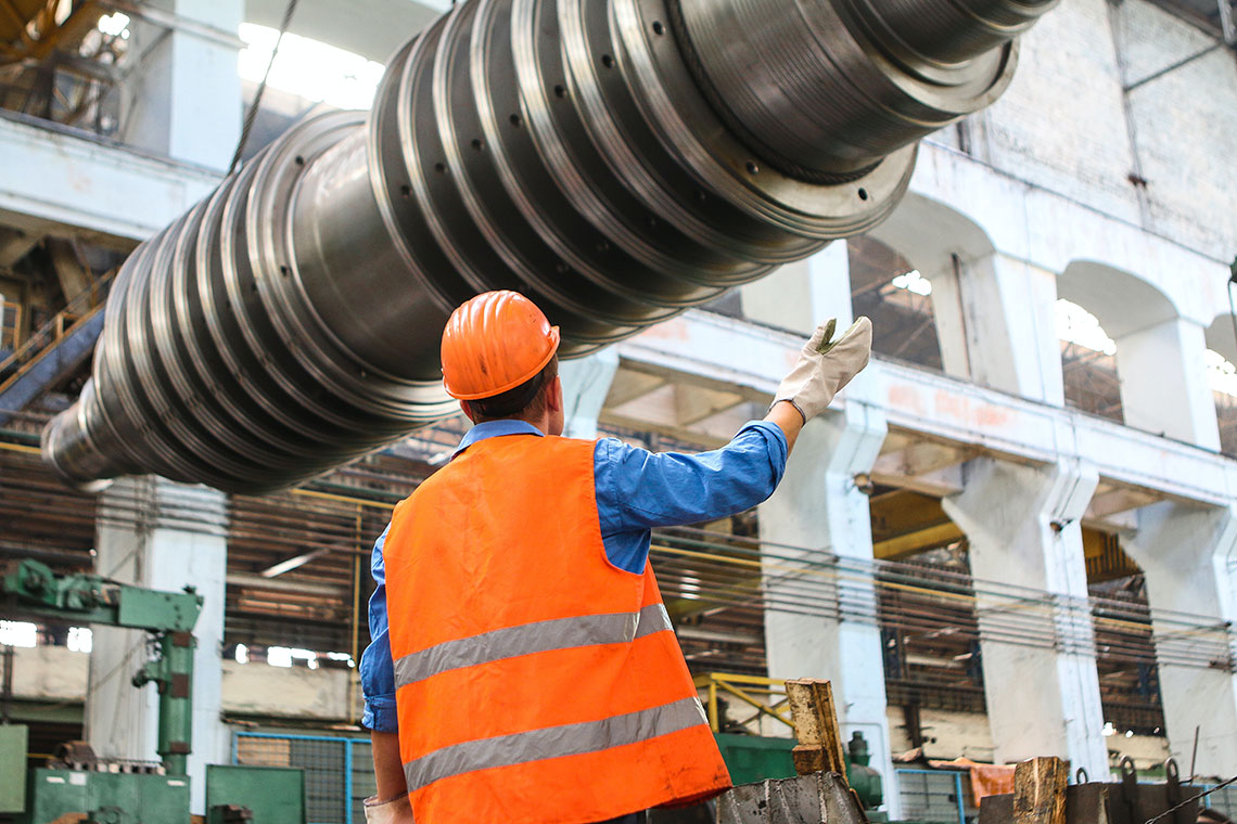 man seen from back in orange hat and vest, under industrial equipment