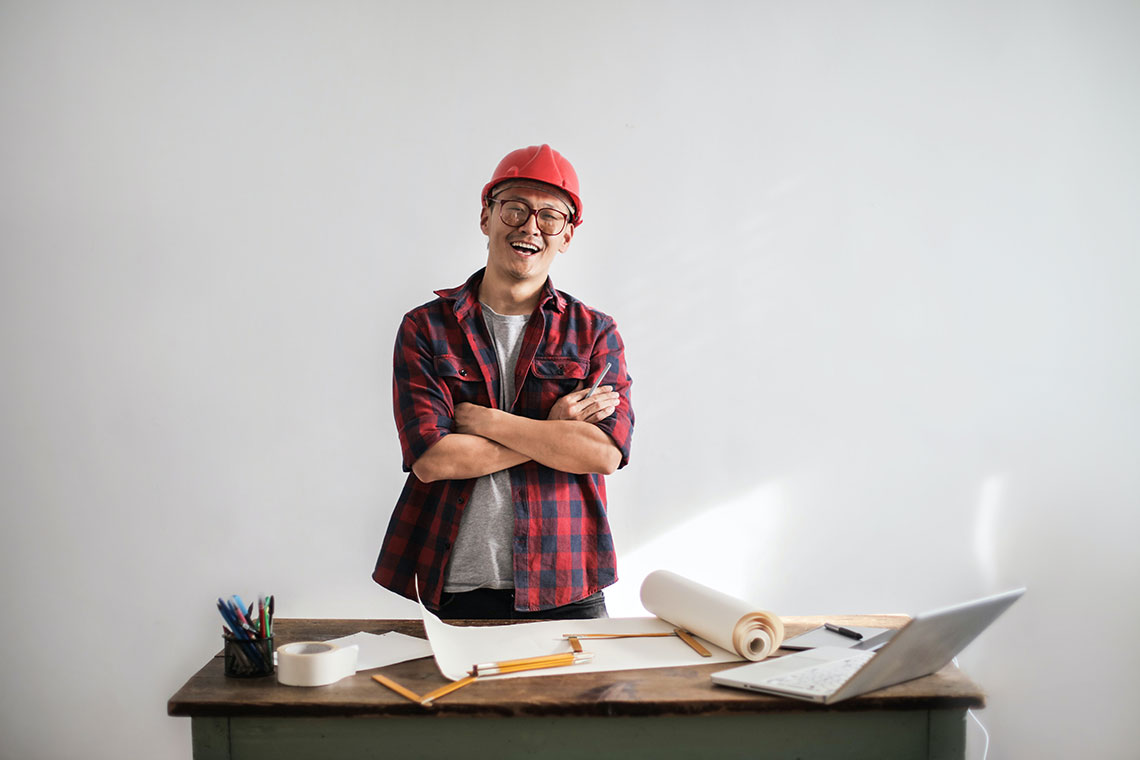 contractor in a hard hat, plaid, smiling behind a desk with blueprint unfurled