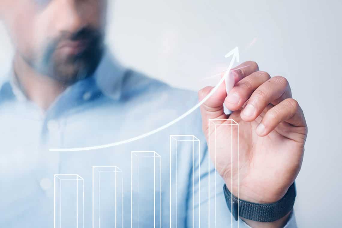 A businessman in a light blue shirt draws a rising bar chart and trend line on a transparent glass board