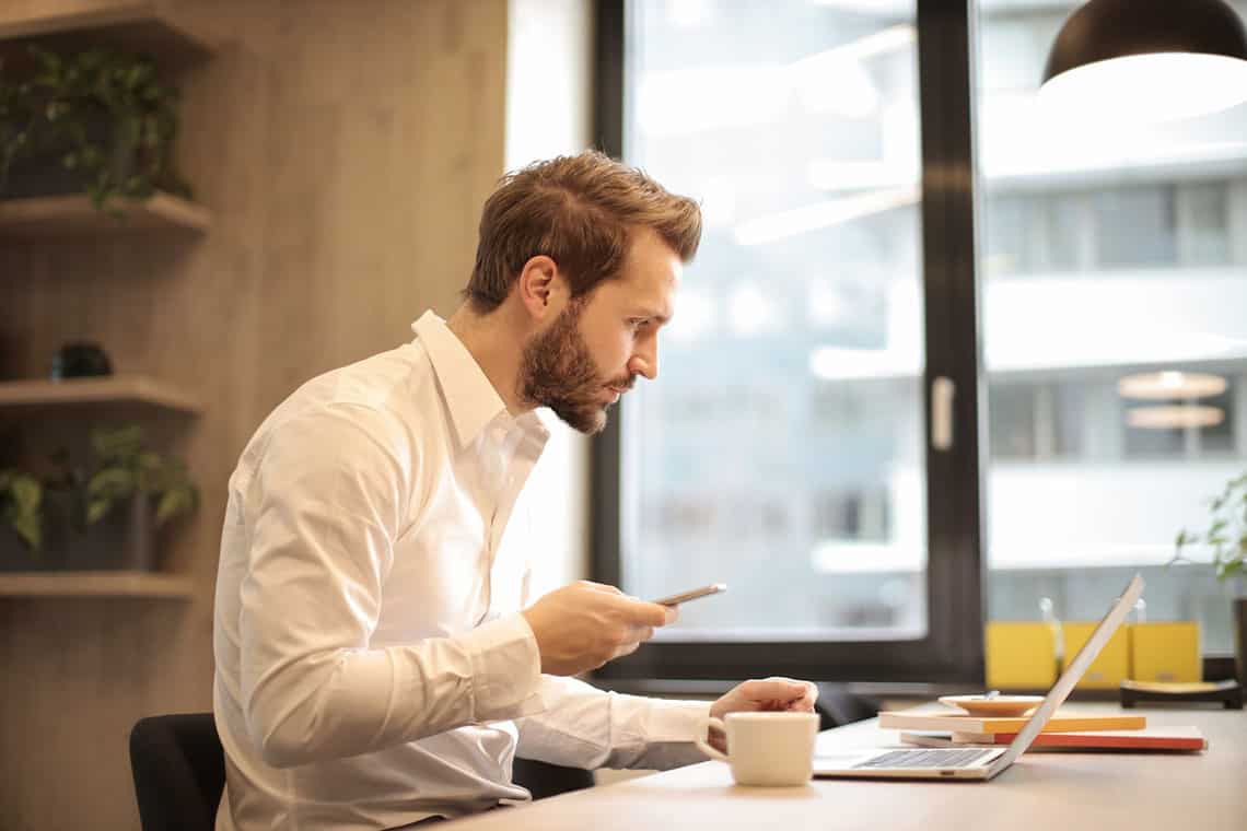 A man in a white shirt sits at a desk, looking at his laptop while holding a smartphone in a bright office