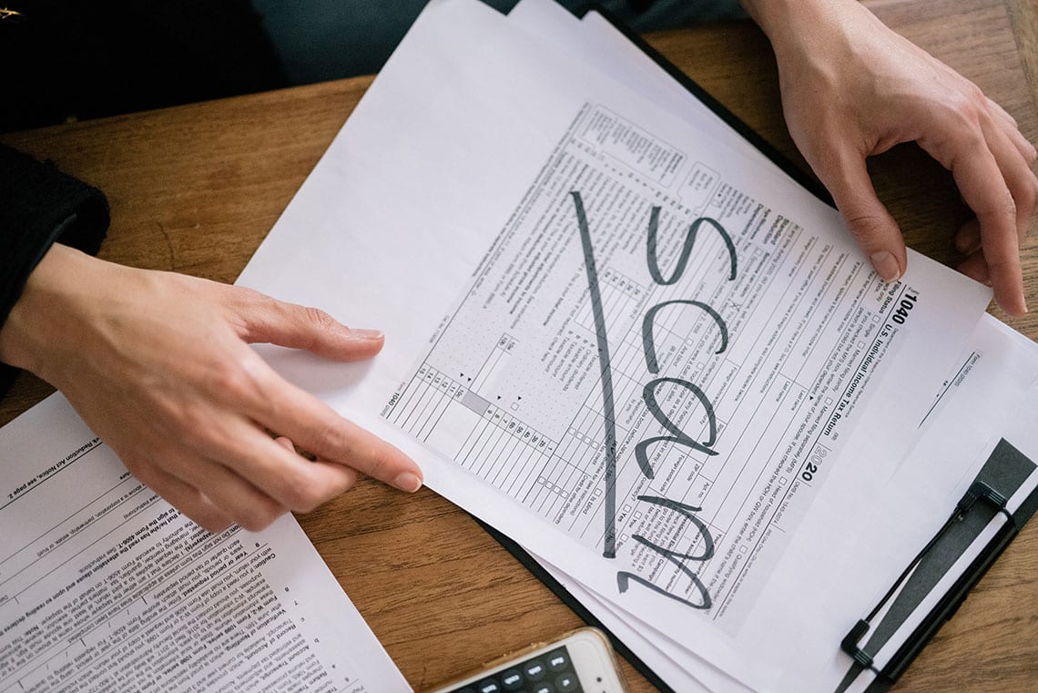 A person holds tax documents on a wooden desk with the word scam written diagonally across the paper