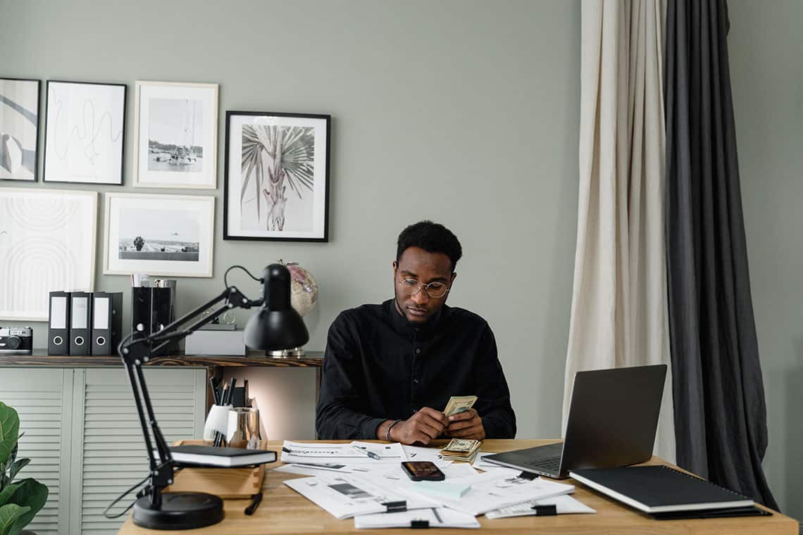 A man in an office counting cash at a cluttered wooden desk with a laptop, papers, and a black lamp