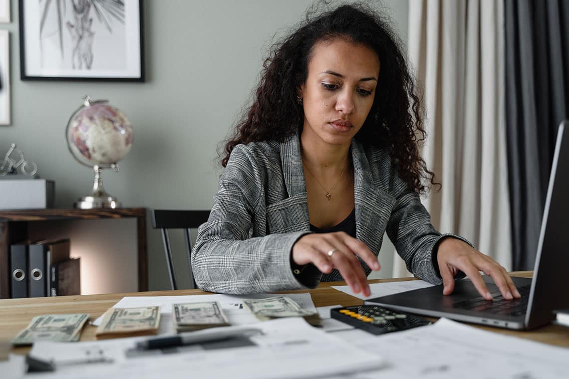 A woman working at a desk with a laptop, calculator, paperwork and stacks of dollar bills